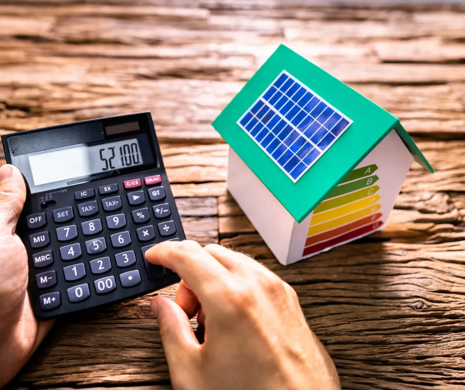 a hand using a calculator next to a small house computing solar panel roofing cost